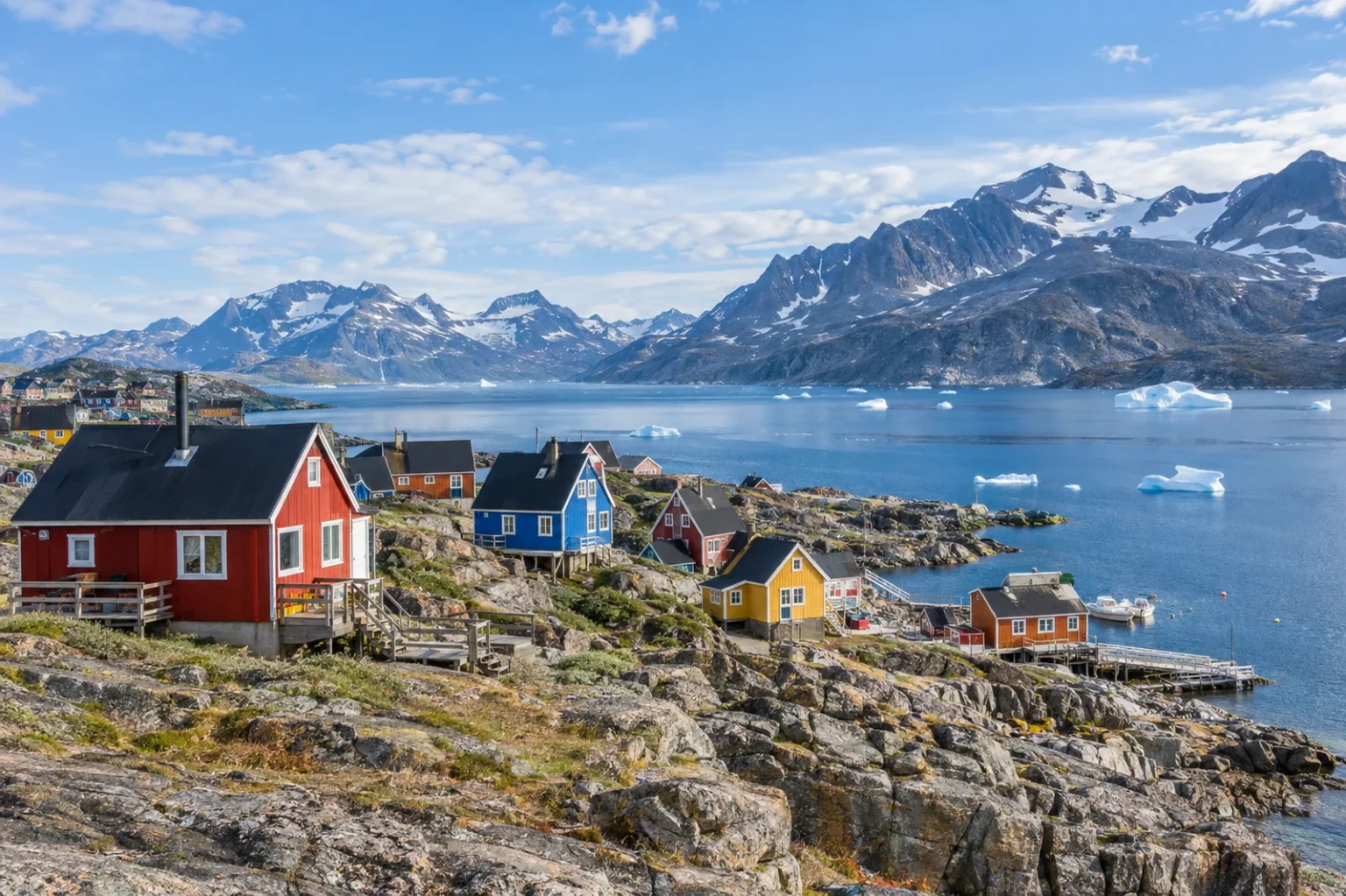 Petites maisons colorées d'un village côtier du Groenland, adossées à la roche, avec des icebergs dans le fjord et des montagnes enneigées en arrière-plan